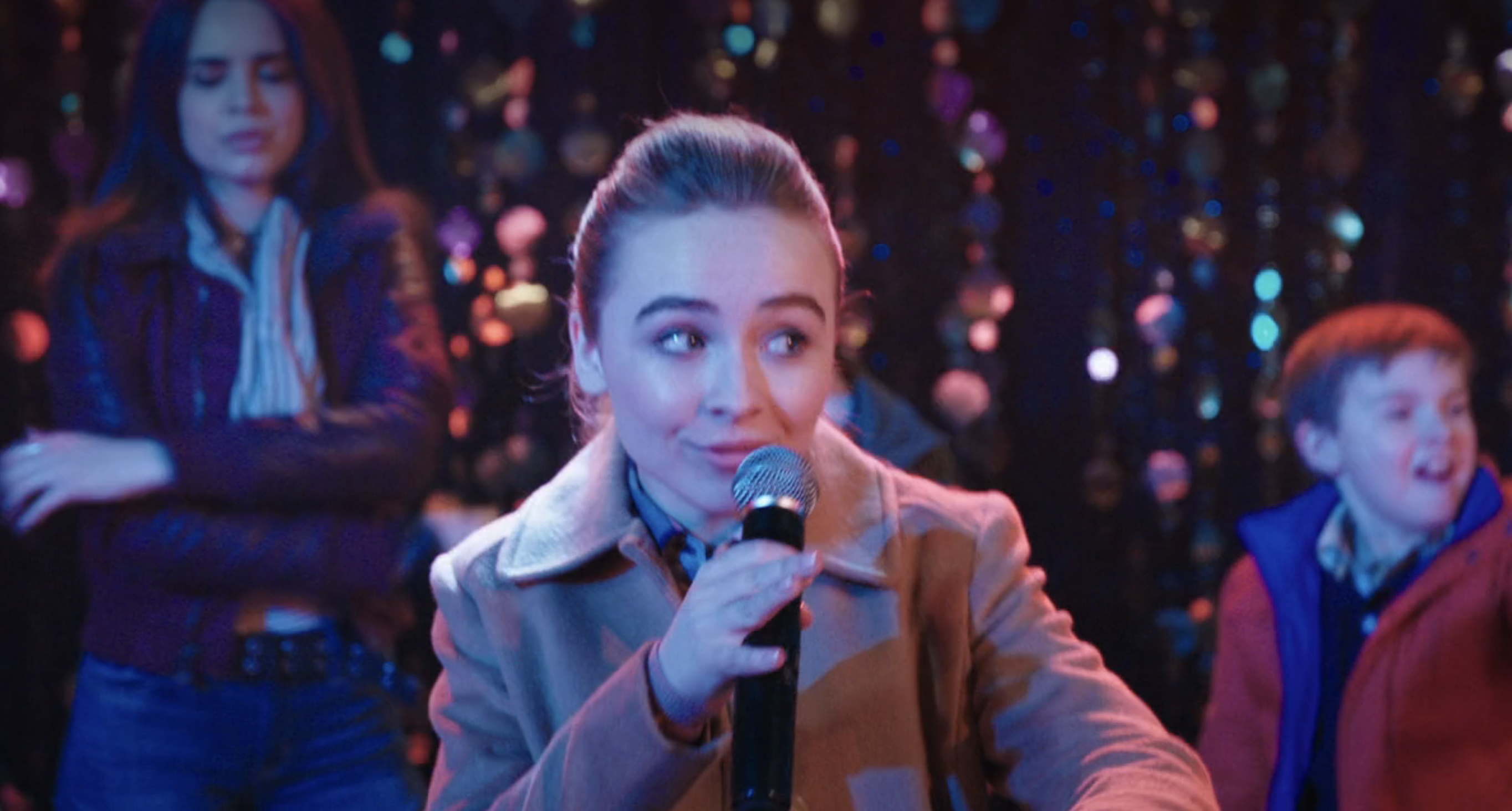 in a scene, Sabrina rapping into a microphone with a crowd in the background