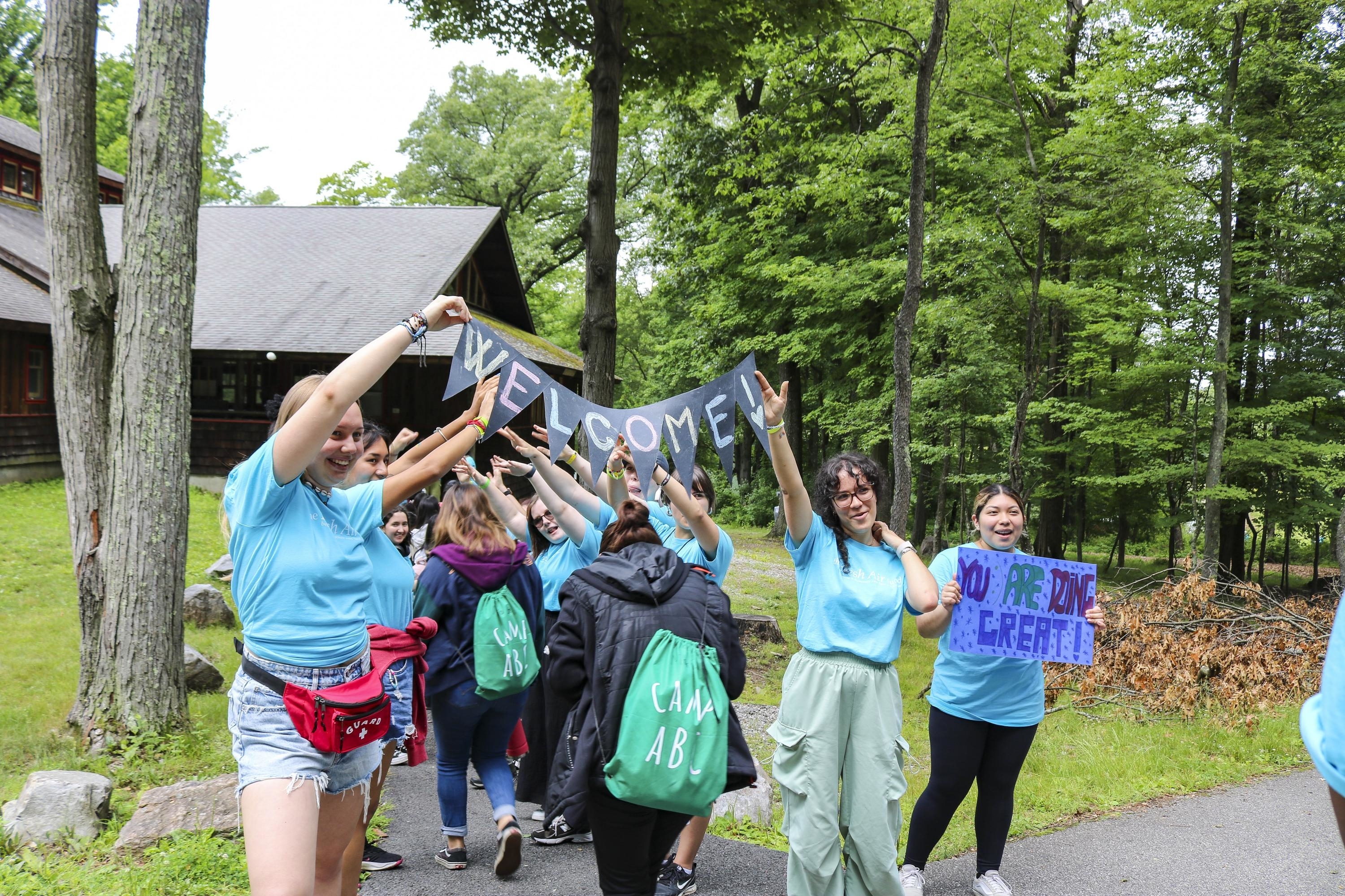 Group of people holding a &quot;WELCOME&quot; banner and cheering outdoors near trees and a building