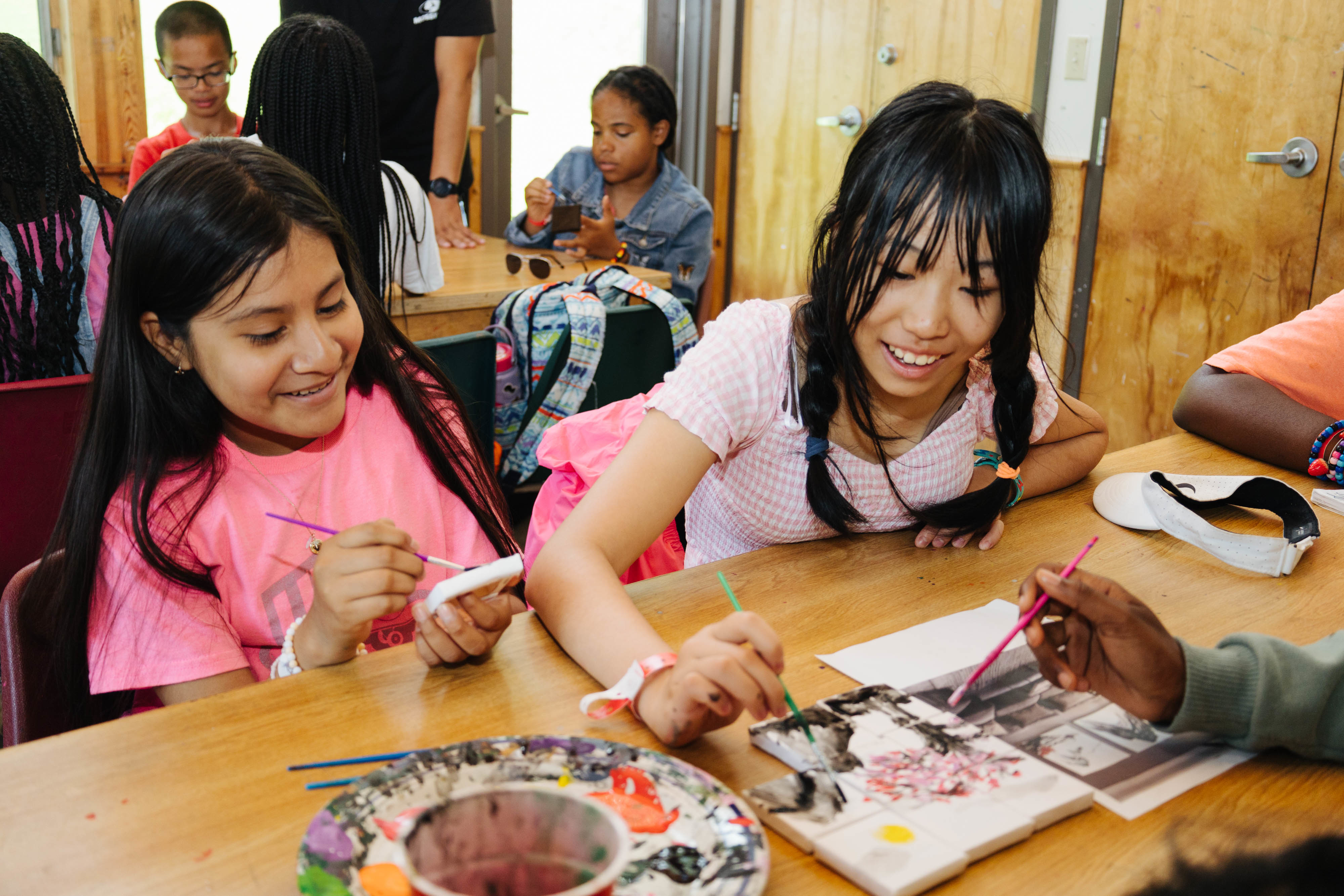 Two girls happily engaging in a painting activity at a table with other children in the background