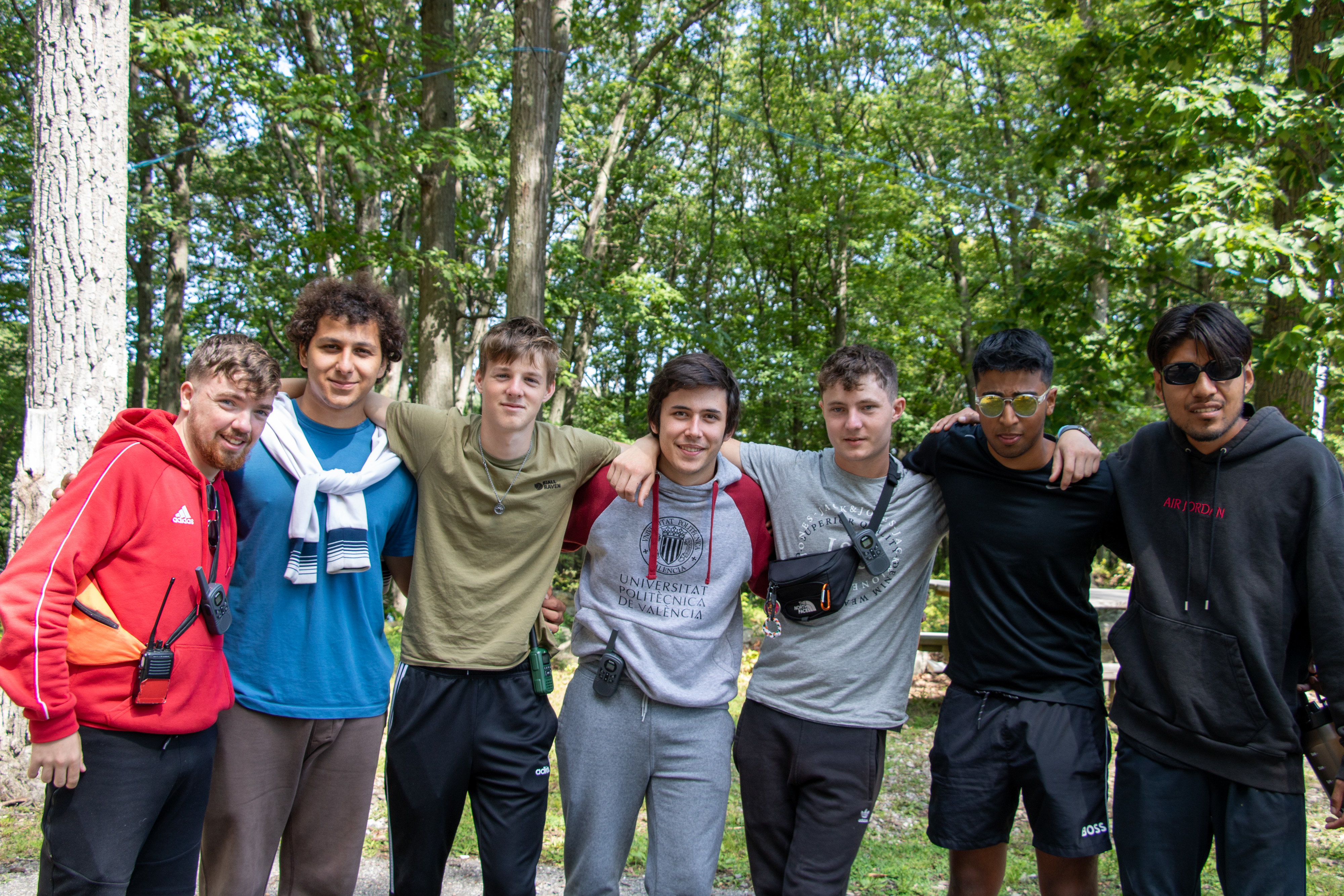 Group of seven friends arm in arm standing outdoors with trees in the background