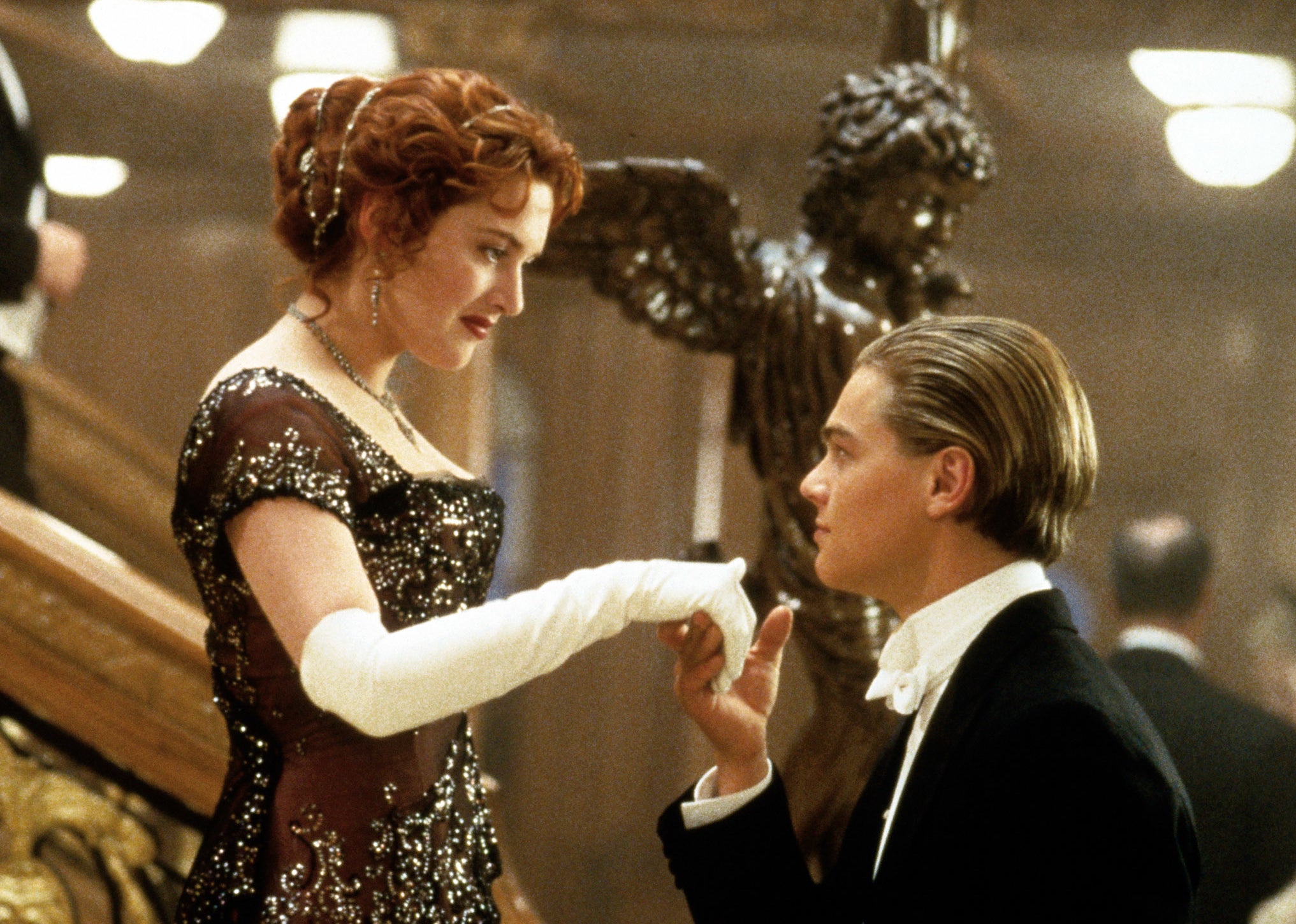 A man in a tuxedo kisses the hand of a woman in an embellished gown on the Titanic&#x27;s grand staircase