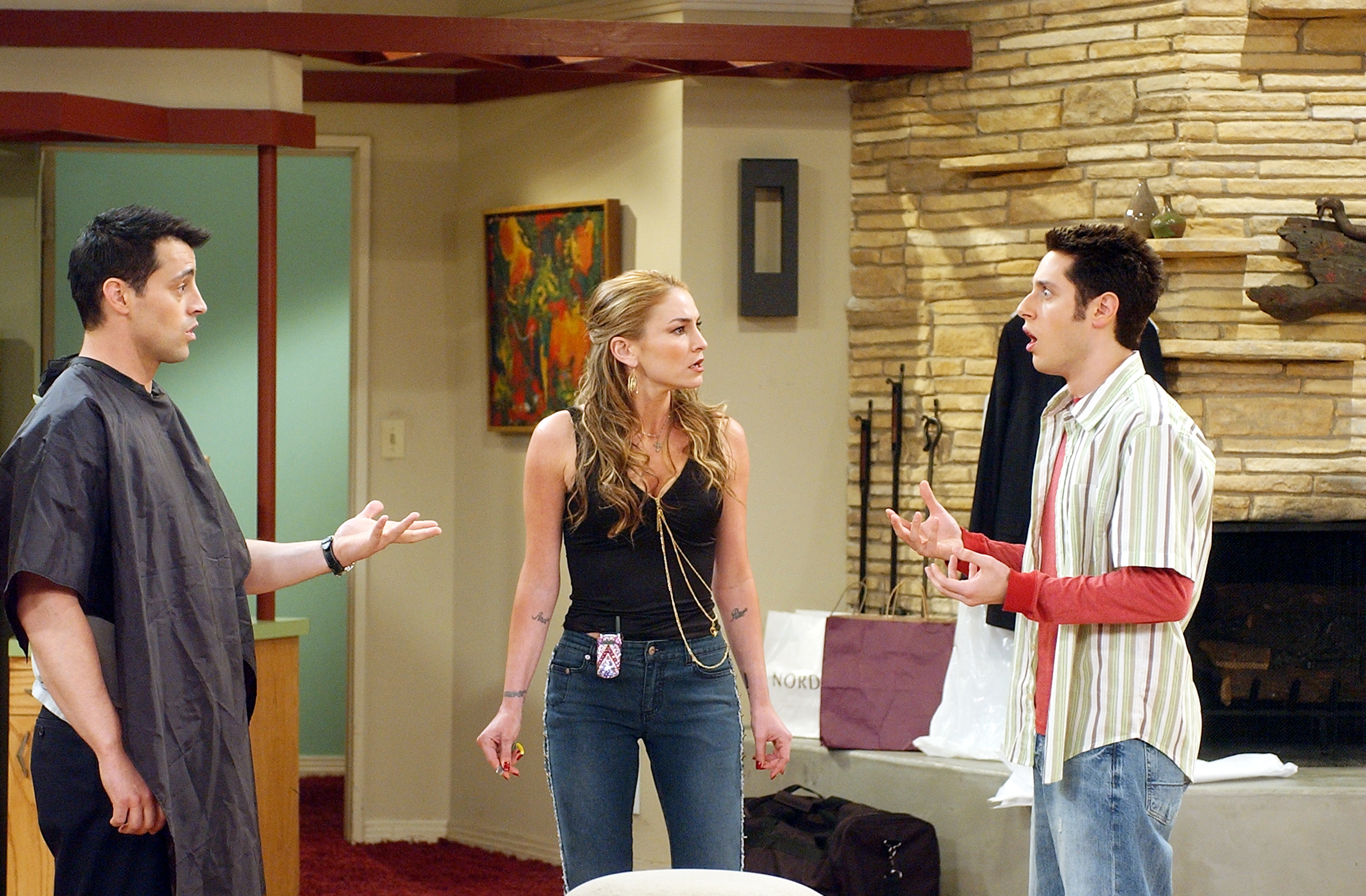 Three people in a sitcom scene with two men gesturing in conversation and a woman in the center looking perplexed