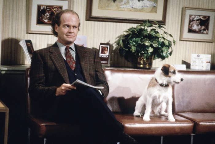 Man in suit sitting with dog on a couch; set resembles a talk show with photos in background
