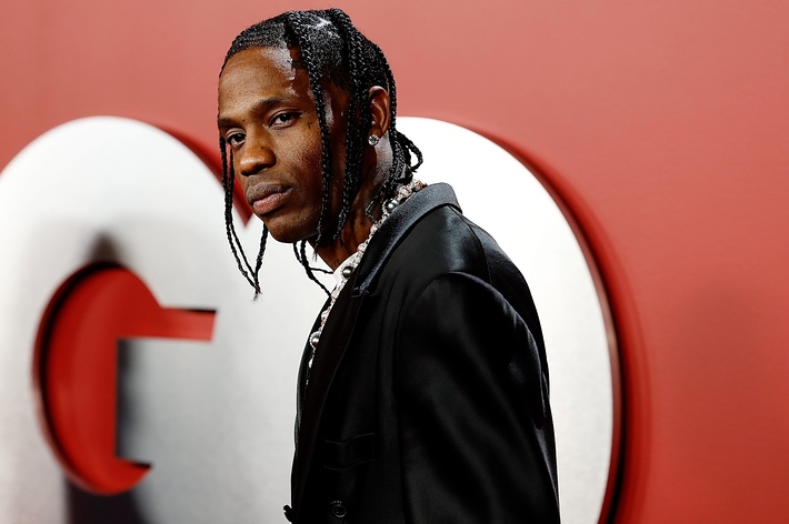 Travis Scott in a black suit with white shirt, standing in front of a large letter "G."