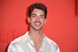 Manu Rios in white shirt smiles at event with red backdrop