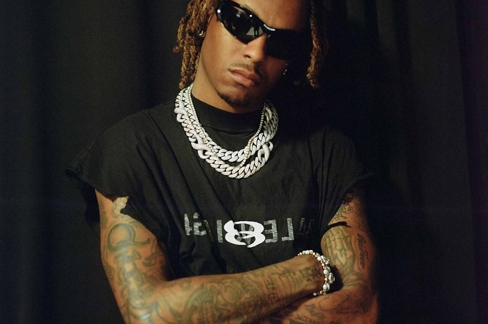 Music artist posing confidently with arms crossed, wearing sunglasses, a chain necklace, and a graphic tee
