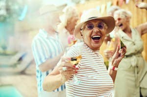 Elderly woman with a wide smile enjoying a party outdoors with friends, wearing a hat and sunglasses