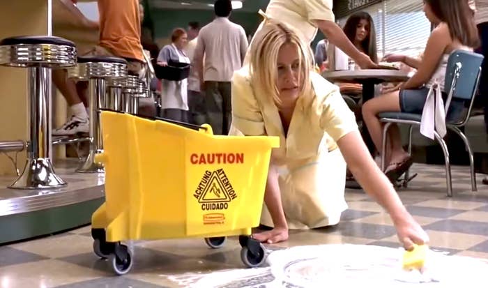 Woman in a yellow uniform cleaning a spill on a diner floor with a caution sign nearby
