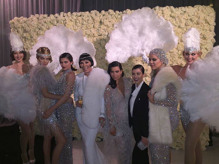 Seven individuals at an event, adorned in sparkly, vintage-inspired attire with feathers, posing in front of a floral backdrop
