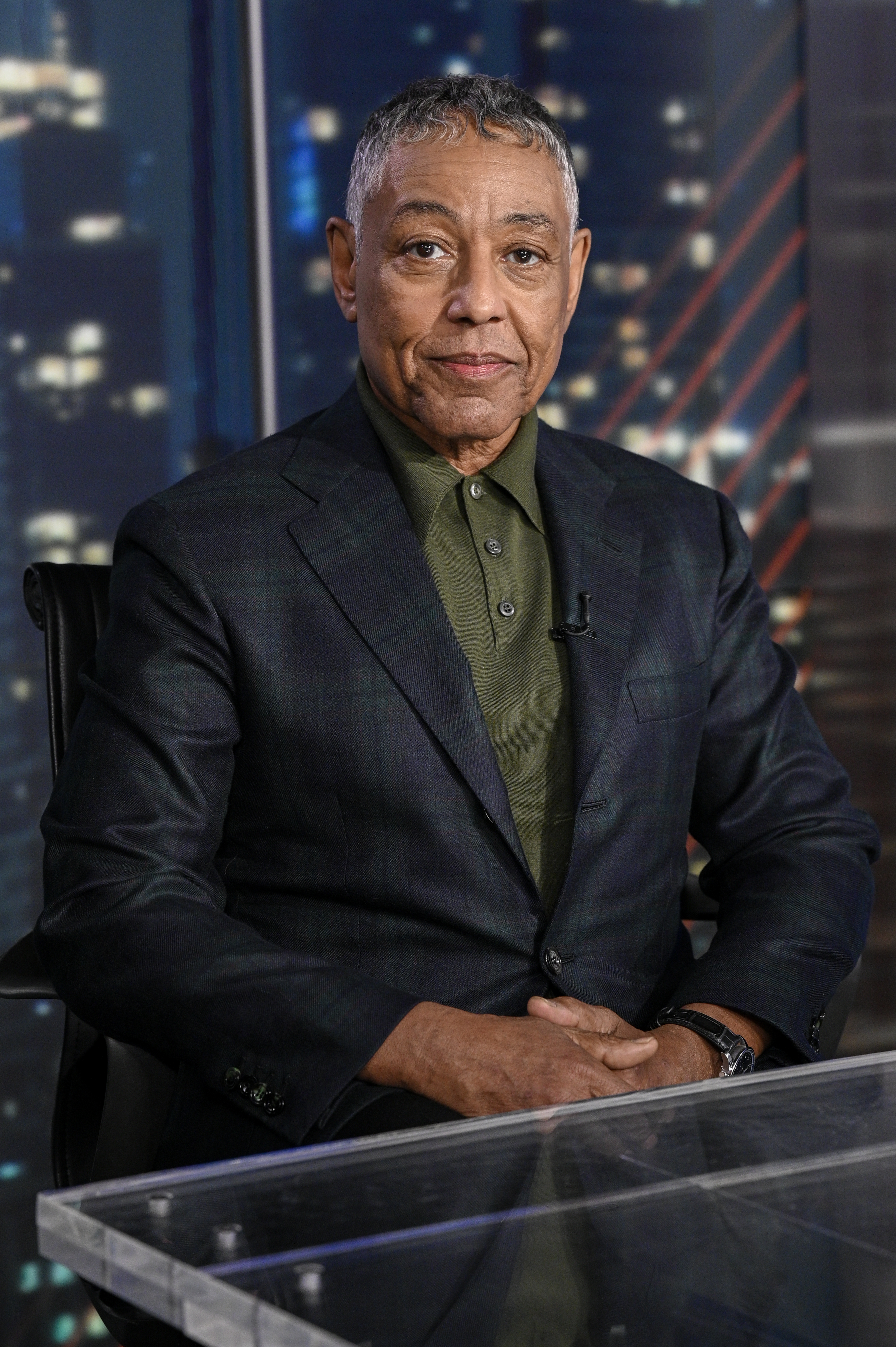 Giancarlo Esposito in a dark suit sitting at a desk, with a blurred cityscape background