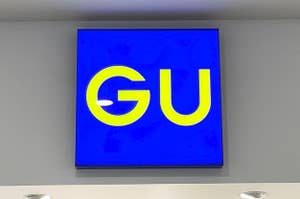 Sign with "GU" logo on blue background, mounted on a wall with ceiling lights visible
