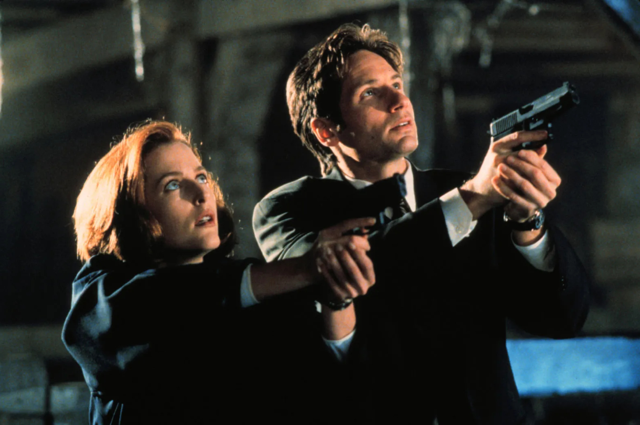 Mulder and Scully from The X-Files aim their guns, standing back-to-back in a tense moment