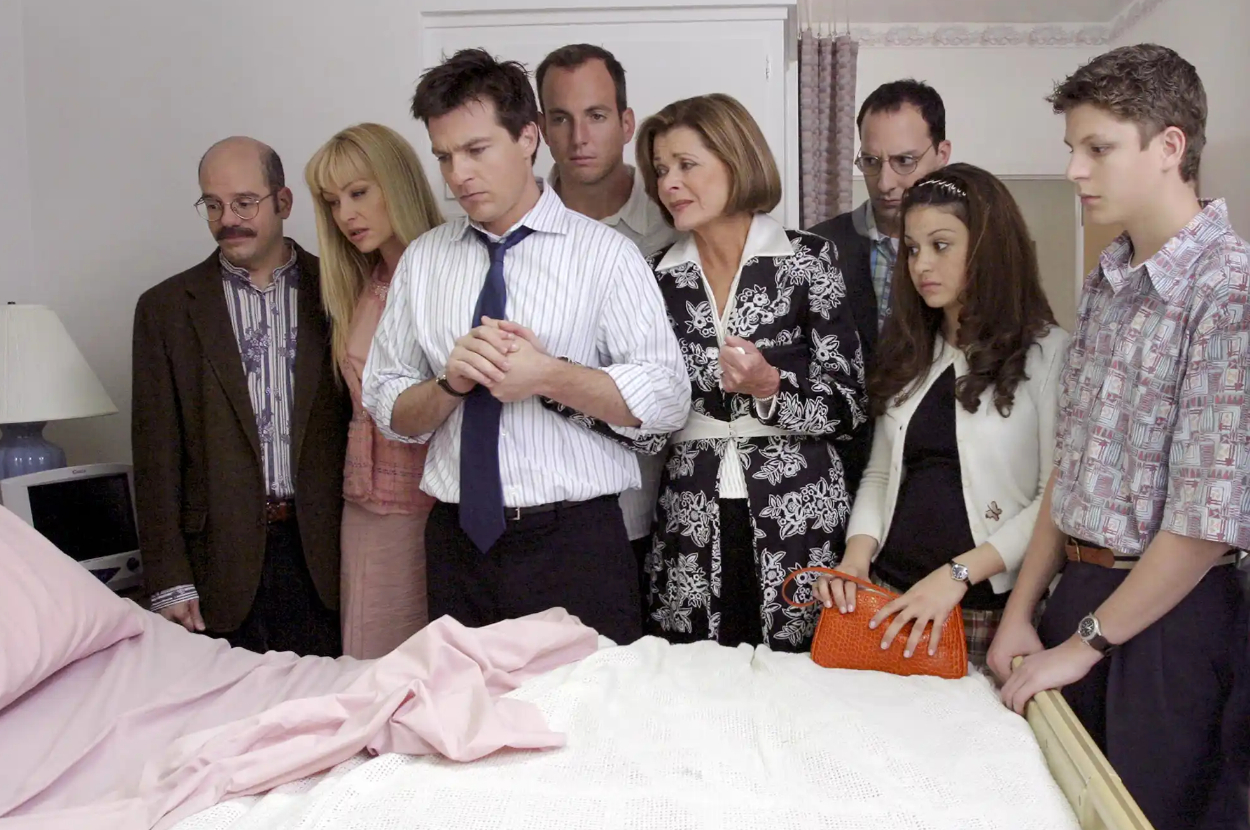 Cast of &quot;Arrested Development&quot; in a scene, standing around a bed with somber expressions