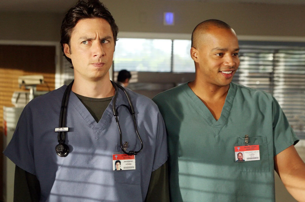 Two actors portraying doctors on a TV show, wearing scrubs with ID badges, standing side by side