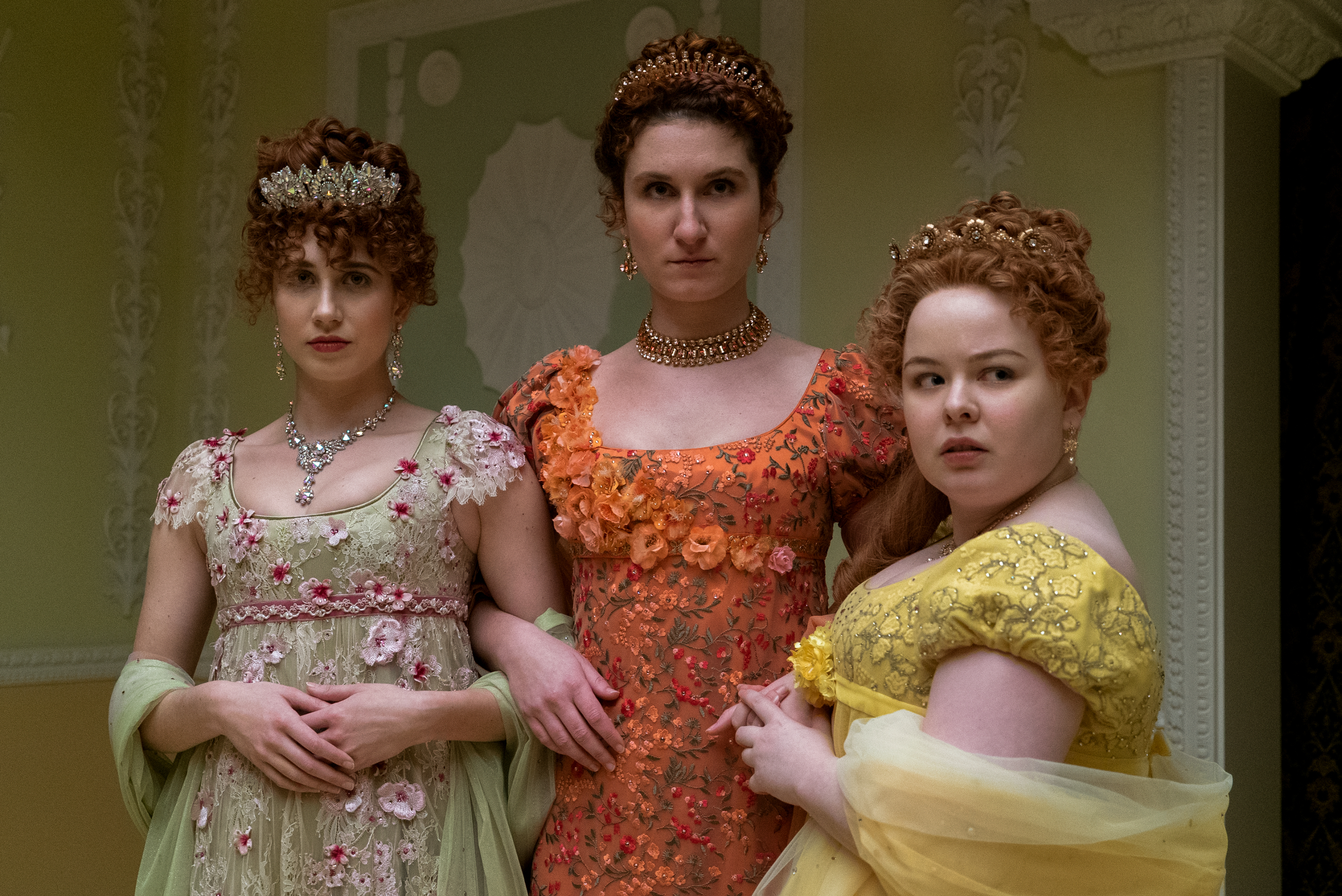 Three characters from Bridgerton in period costumes; one in green with pink flowers, middle in ornate orange, and one in yellow with ruffles
