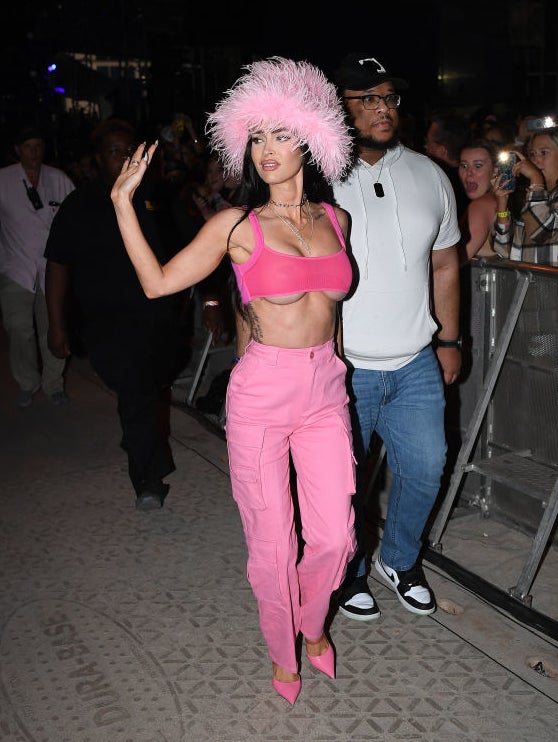 Megan Fox in a pink outfit with a feathered hat, waving, with a bodyguard and fans in the background