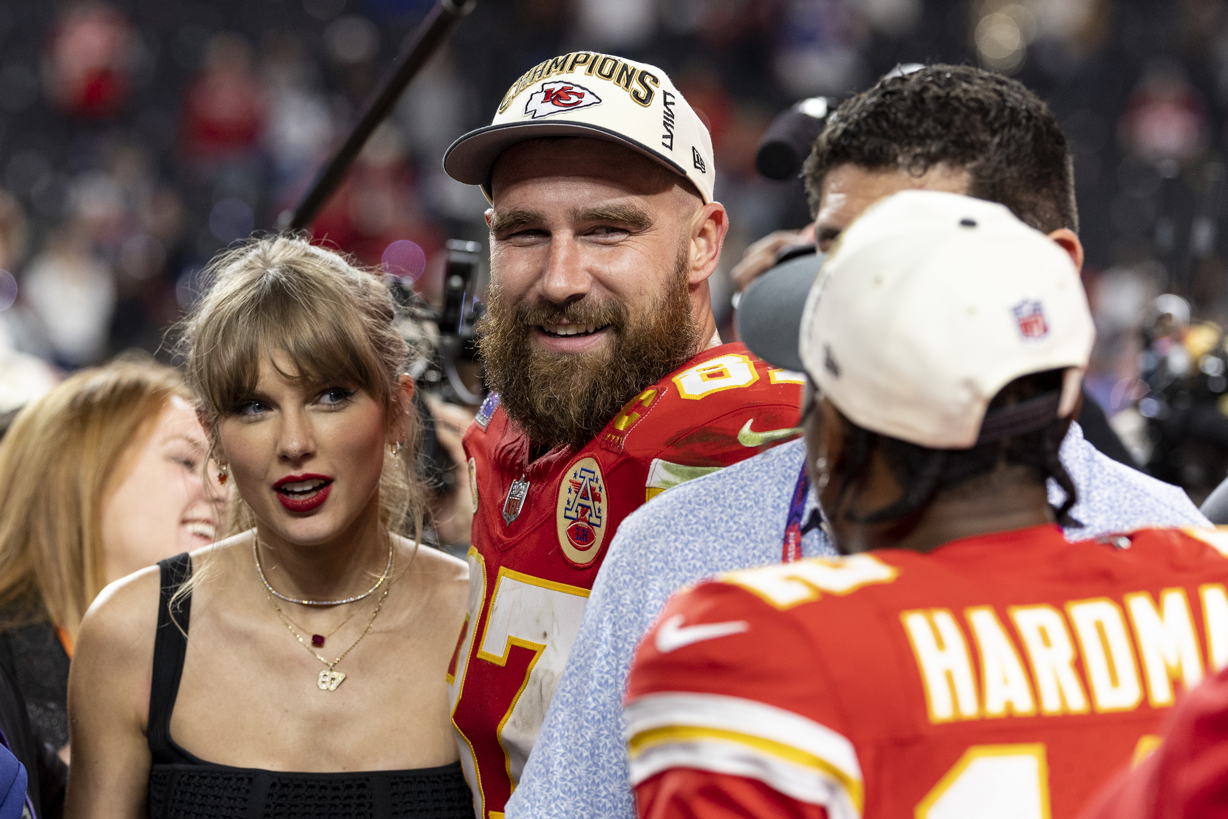 Travis Kelce in a jersey and hat stands next to Taylor Swift after a football game