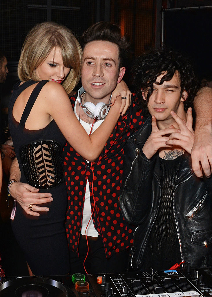 Taylor Swift in a backless dress embracing Nick Grimshaw, who stands next to Matt Healy at an event