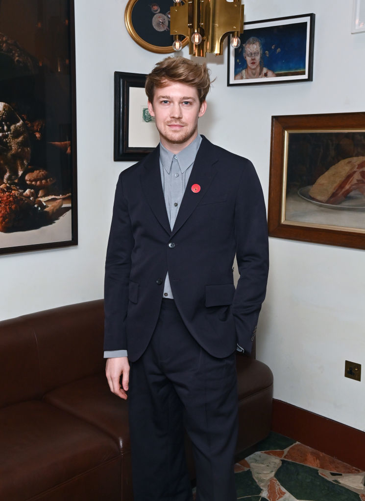 Joe in a suit with a pin stands in a room with artwork and furniture