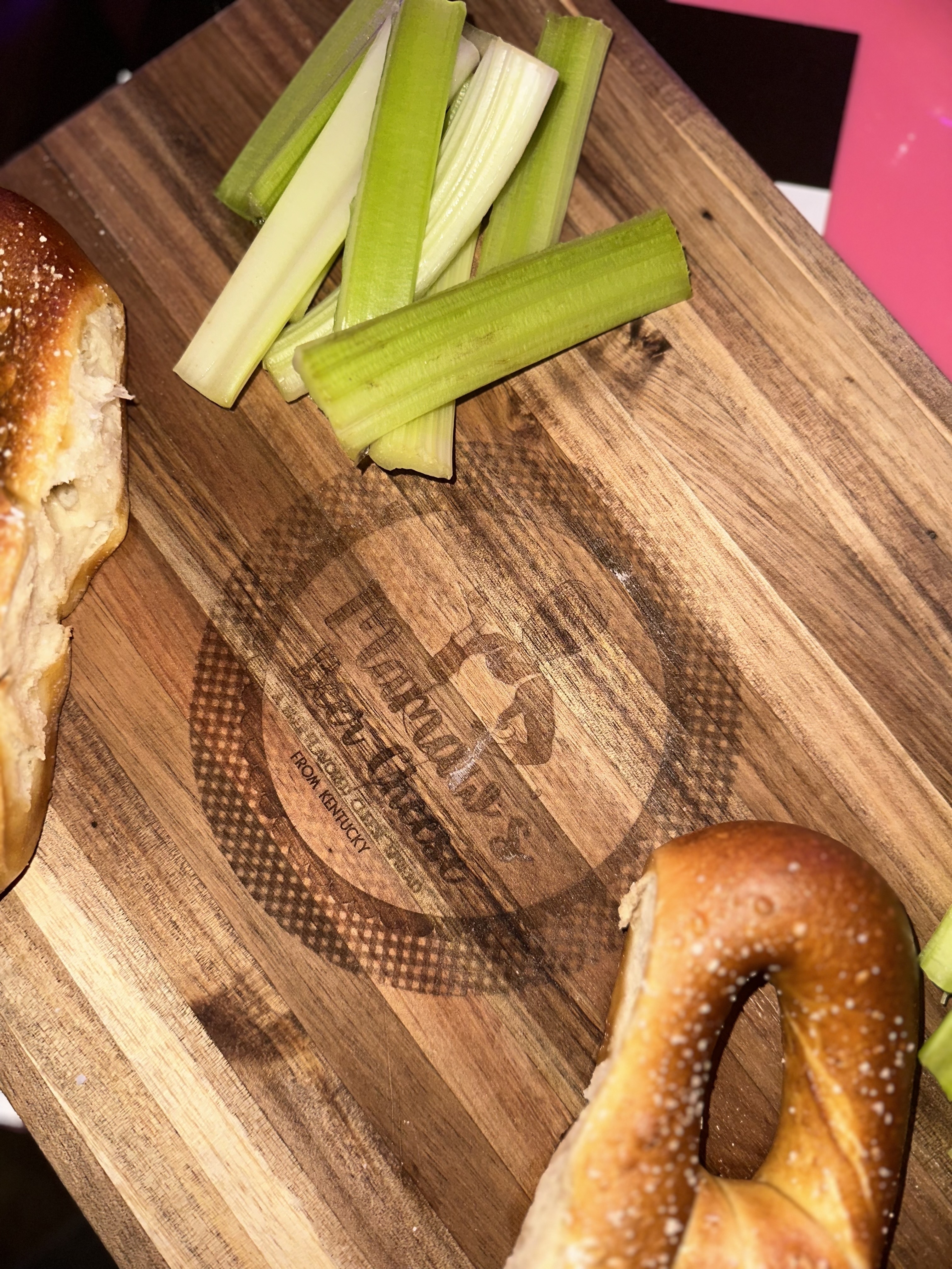 Pretzel and celery sticks presented on a wooden cutting board with an engraved logo