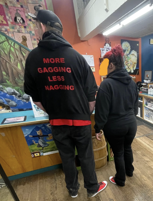 Two people standing in a shop; one has a sweatshirt with &quot;MORE GAGGING LESS NAGGING&quot; text on the back