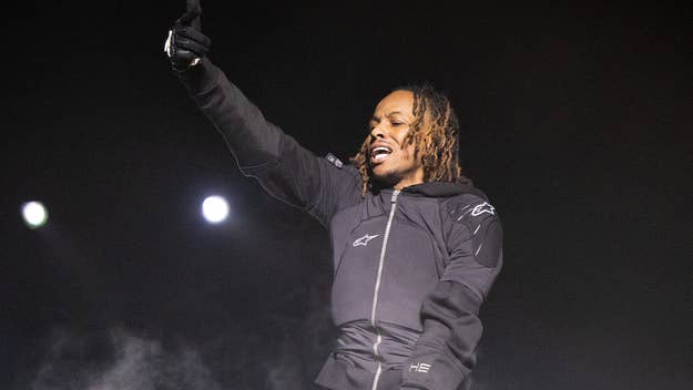 Person on stage performing, wearing a logoed hoodie, gesturing upwards with one hand