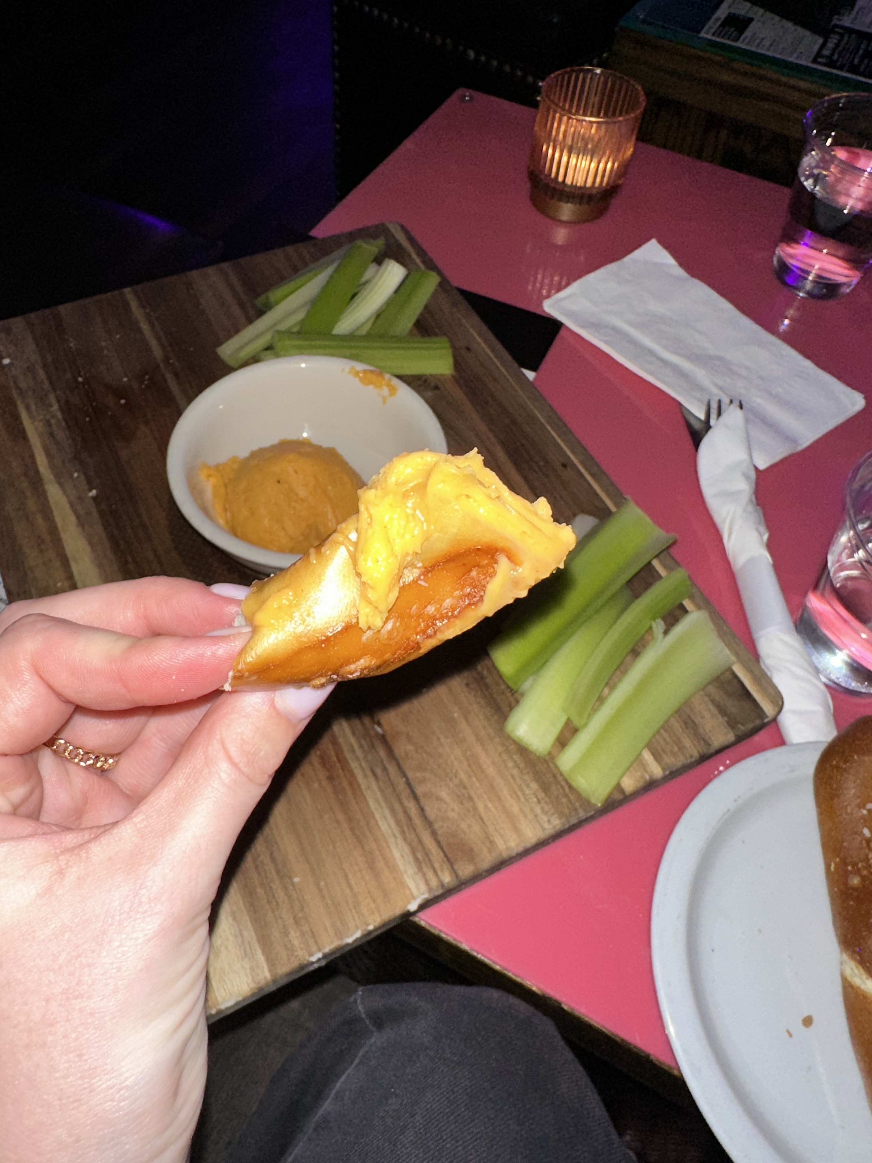 Person&#x27;s hand holds a piece of buffalo chicken wing, with celery sticks and sauce on the side on a table