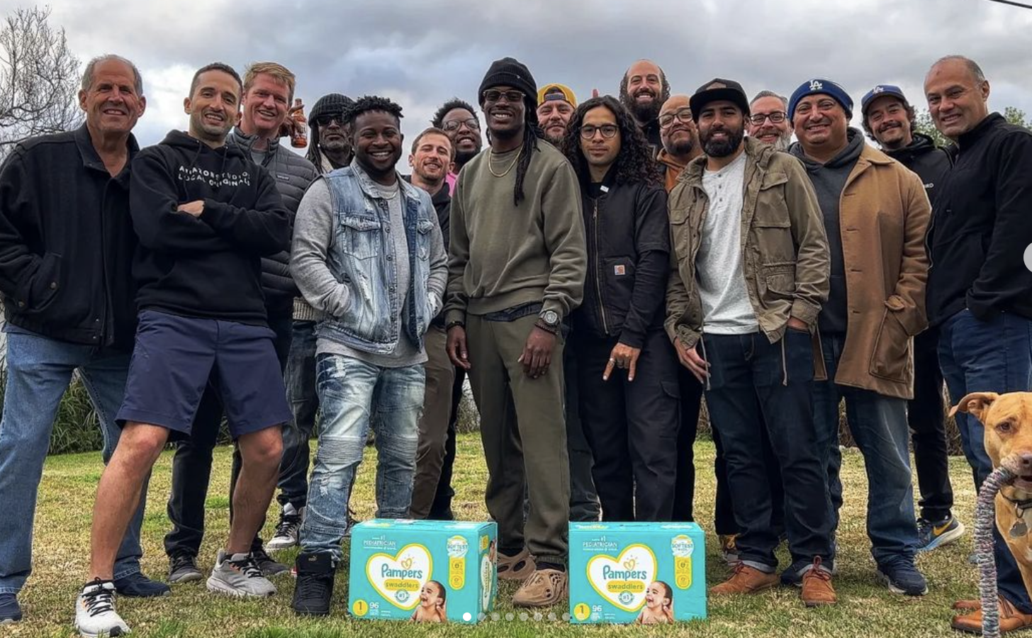 Group of people smiling outdoors with two boxes of Pampers in front, a dog on the right