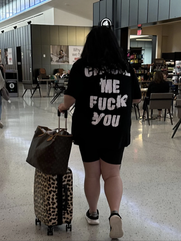Person in a graphic tee with explicit text, pulling a suitcase in an indoor space