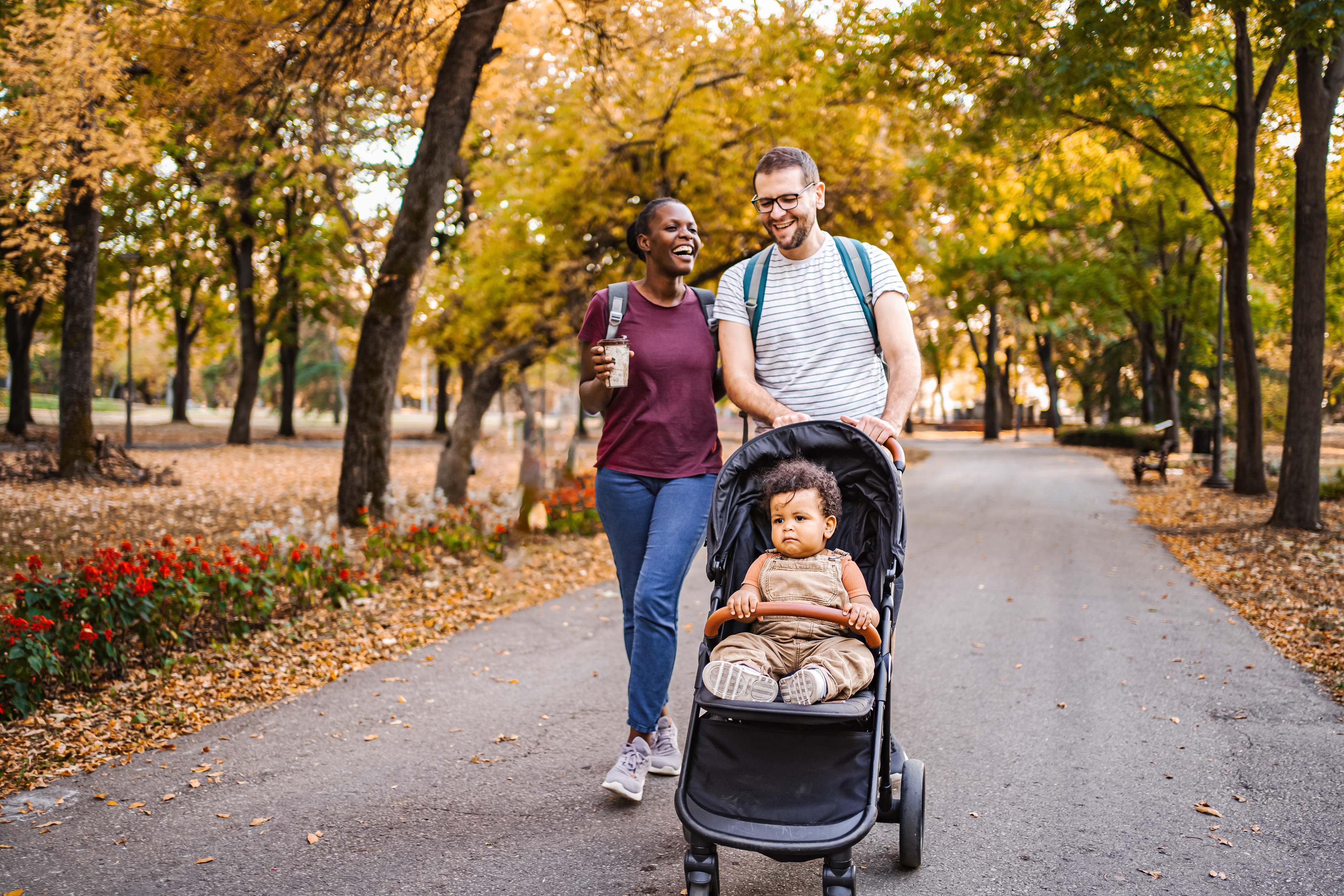 Two adults smiling and walking with a baby in a stroller through a park with trees