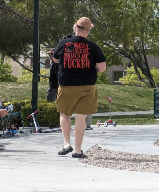 Person in t-shirt and shorts walking at a park, with obscured text on shirt, near children&#x27;s scooters