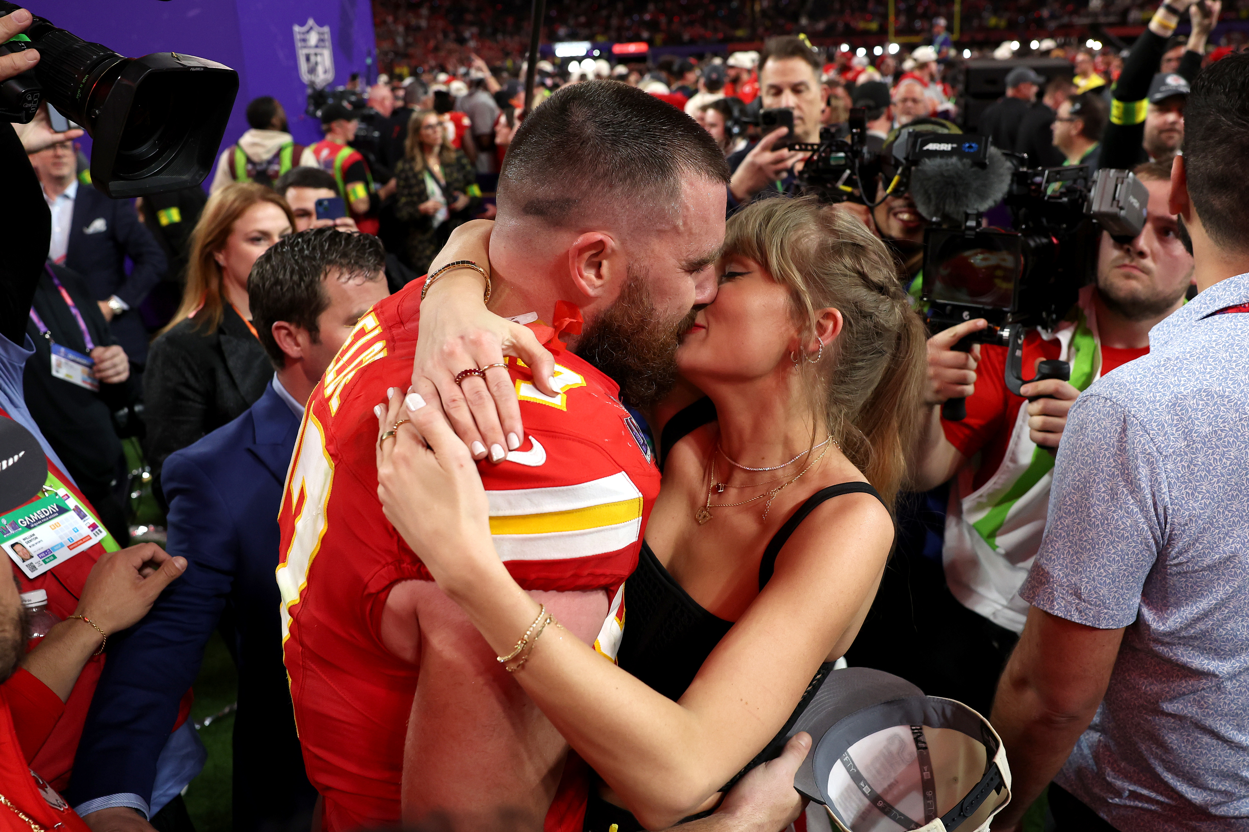 Taylor Swift y Travis Kelce besándose rodeados de fotógrafos después de un evento deportivo