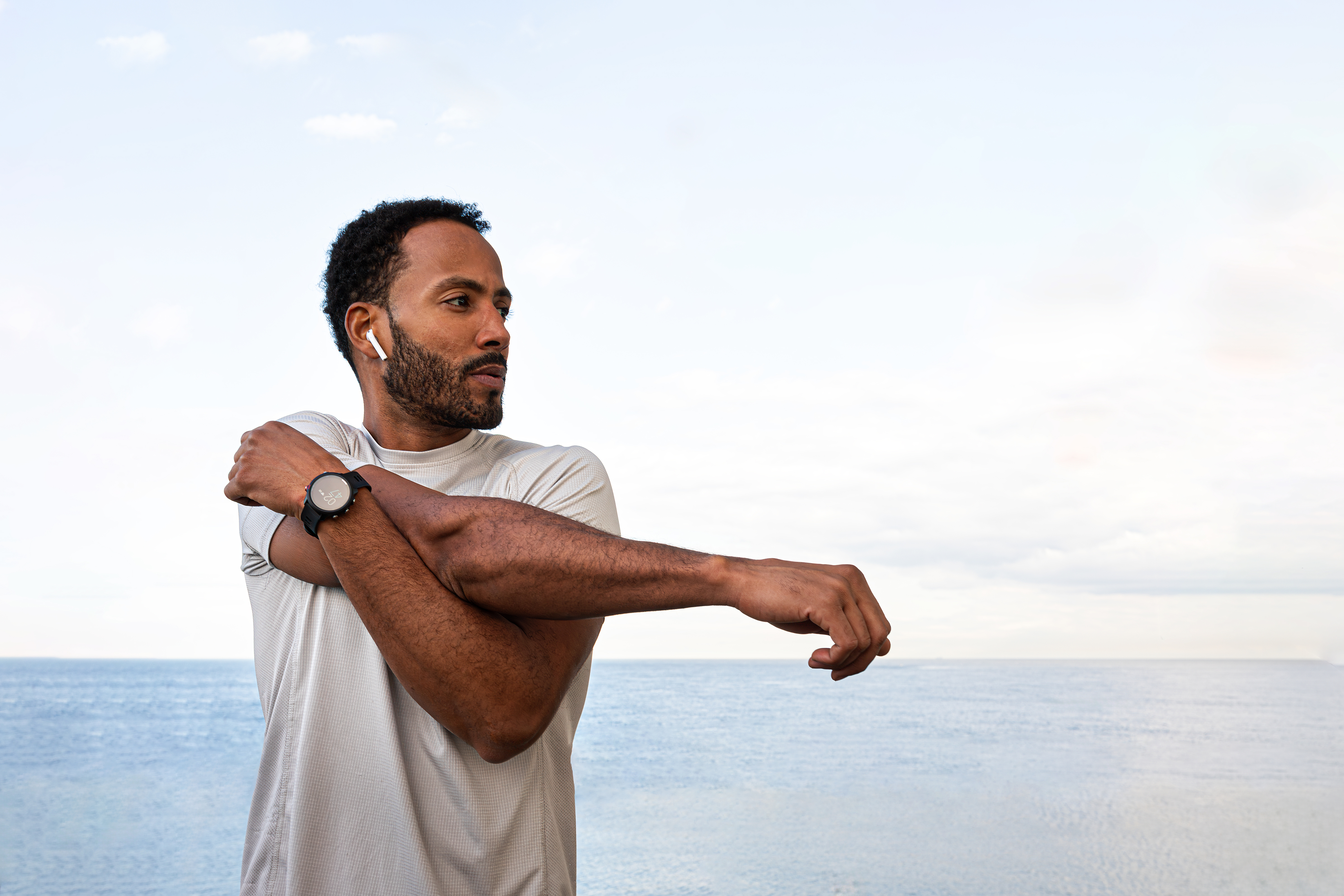 Person stretching their arm across their chest, facing body of water. Wearing casual attire with a watch