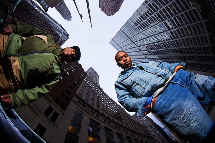 Two men, Nas and DJ Premier, sitting on a curb with skyscrapers rising around them, promoting "Define My Name"
