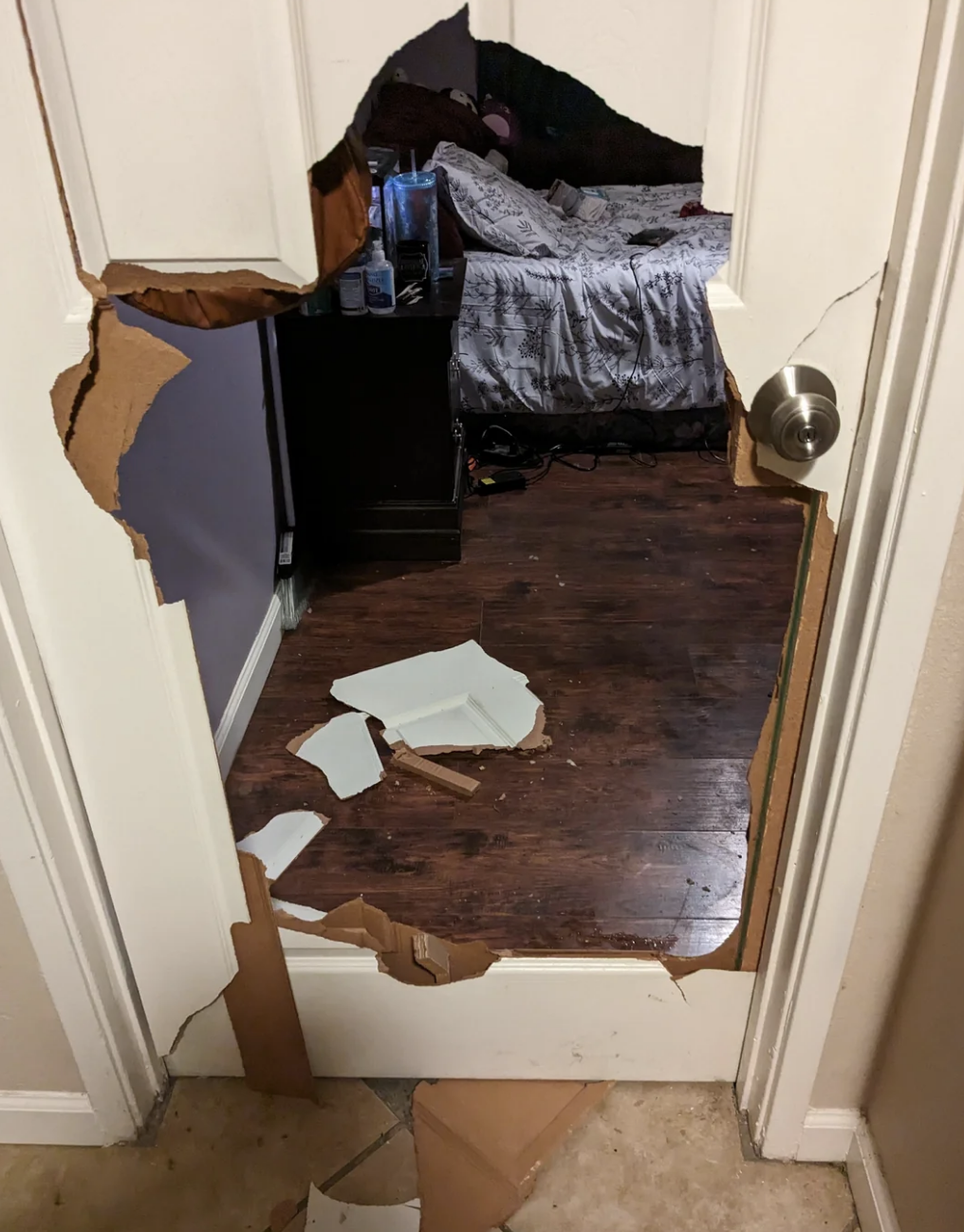 A damaged door with a large hole revealing a room with a bed and scattered debris on the floor