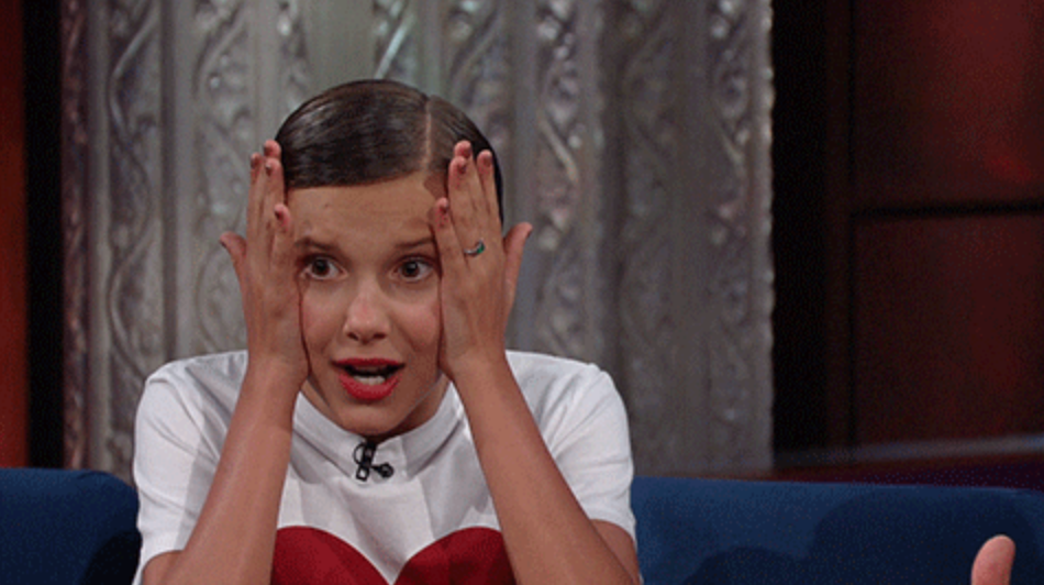 Millie Bobby Brown sitting, hands on face, white top with red heart, surprised expression