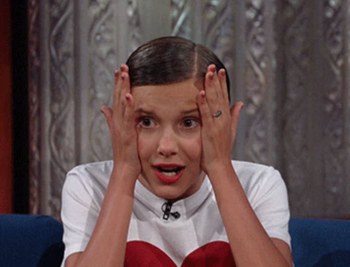 Millie Bobby Brown sitting, hands on face, white top with red heart, surprised expression