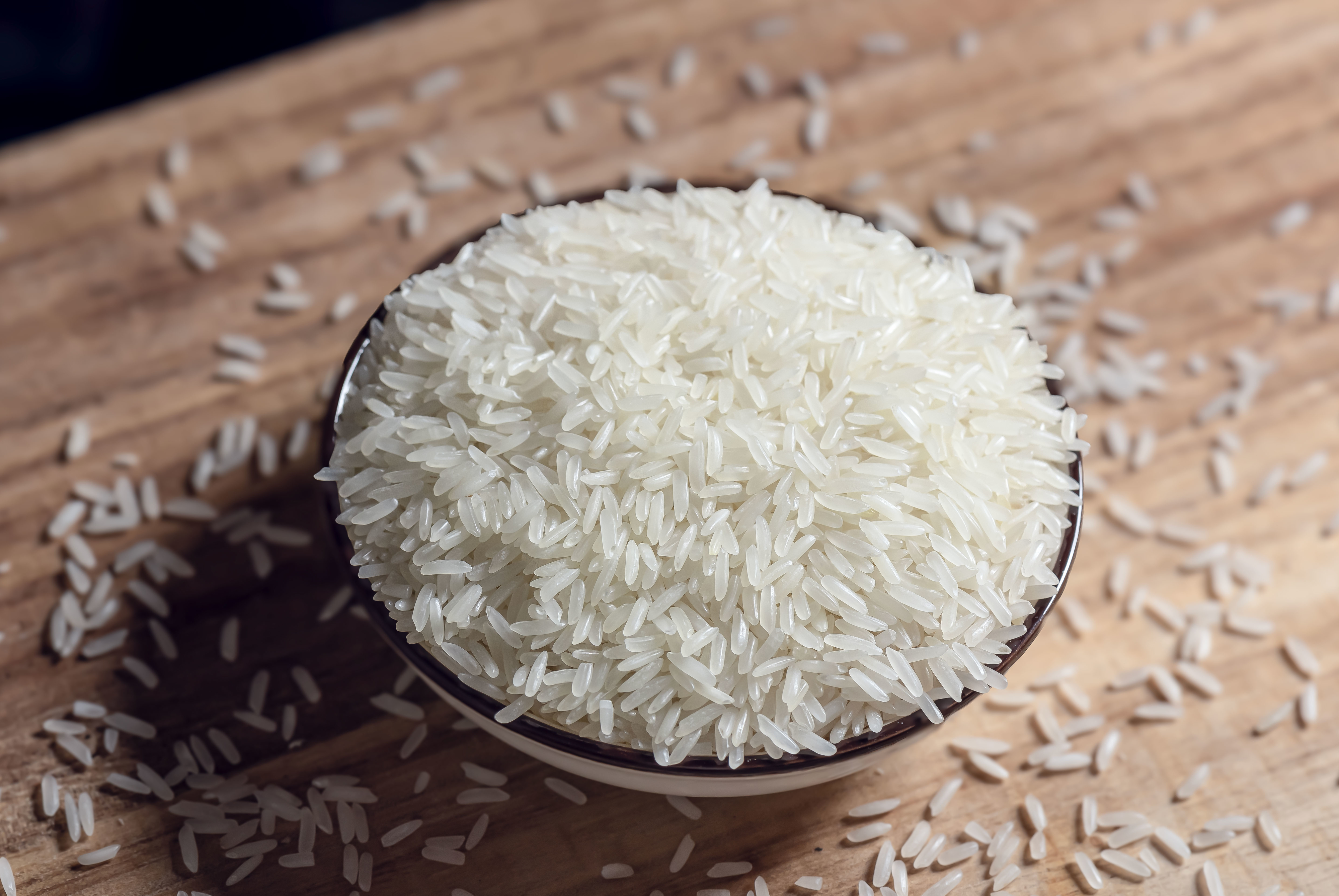 Bowl of uncooked white rice on a wooden surface with grains scattered around it