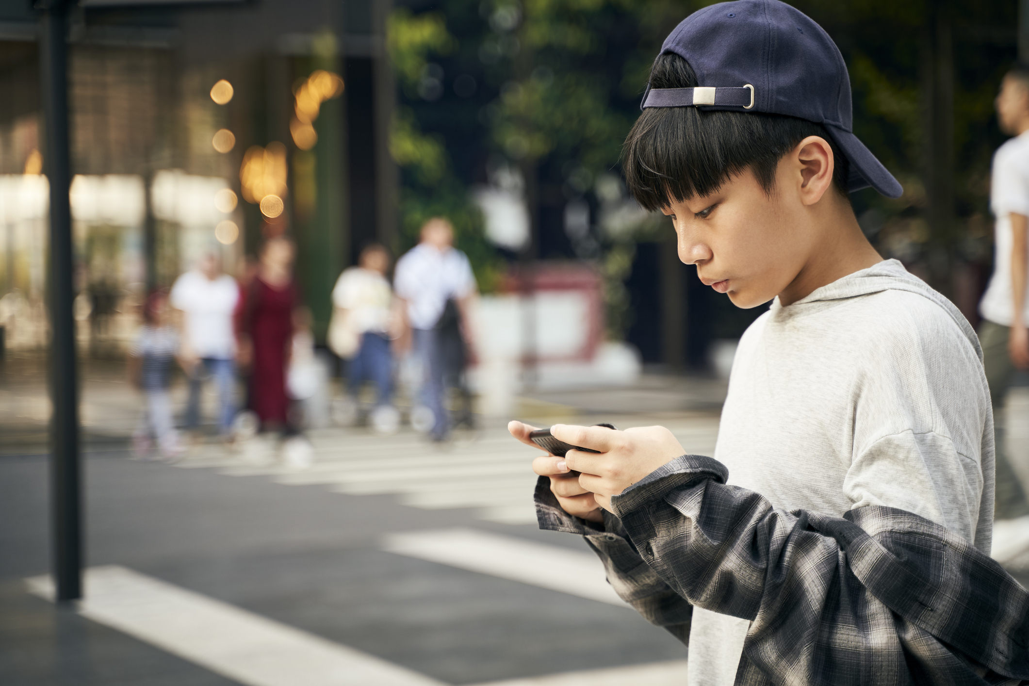 Teen in a cap and layered shirts focused on smartphone with blurred people walking in the background