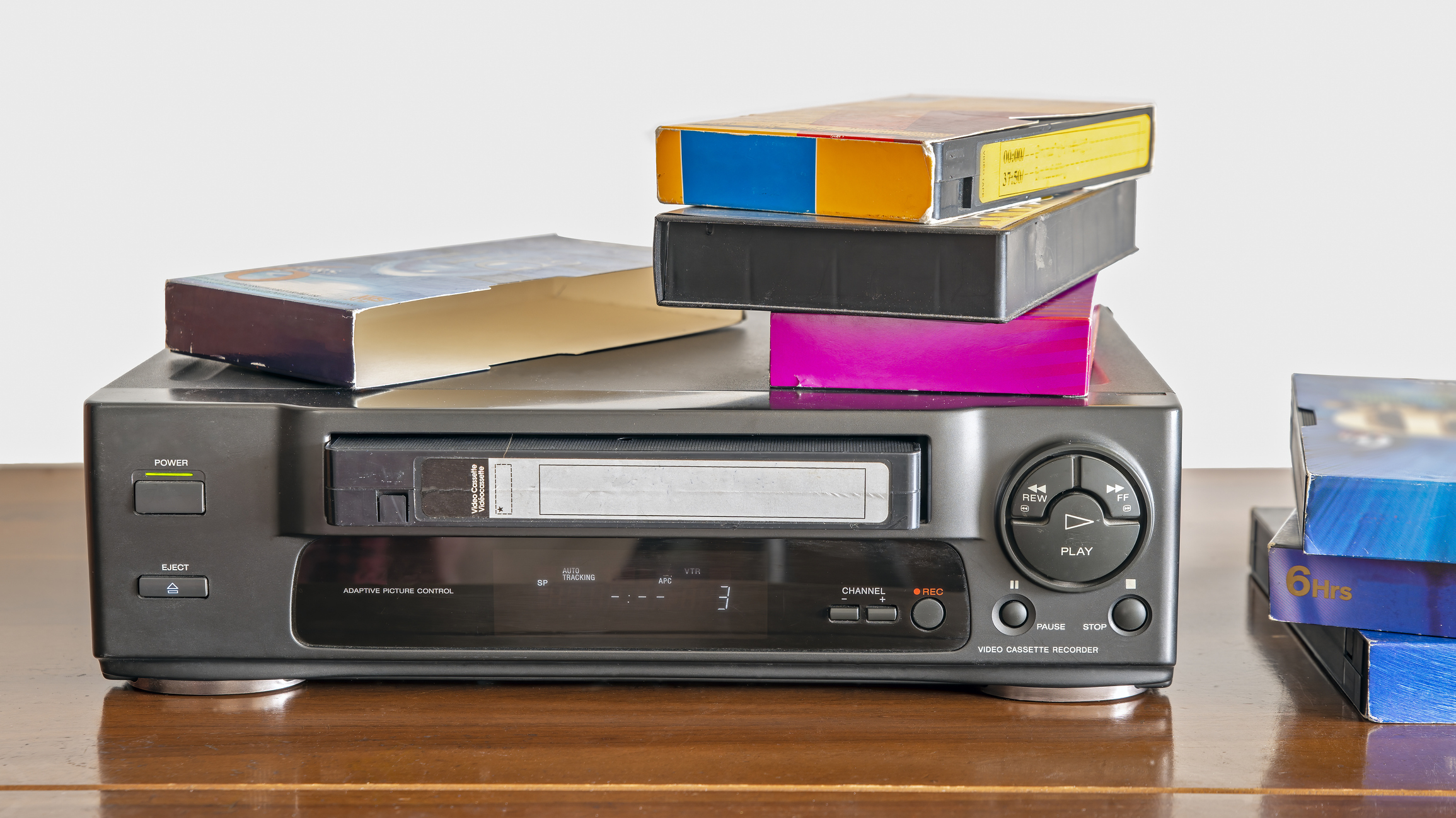 Pile of VHS tapes on top of a VCR with more tapes beside it on a wooden surface