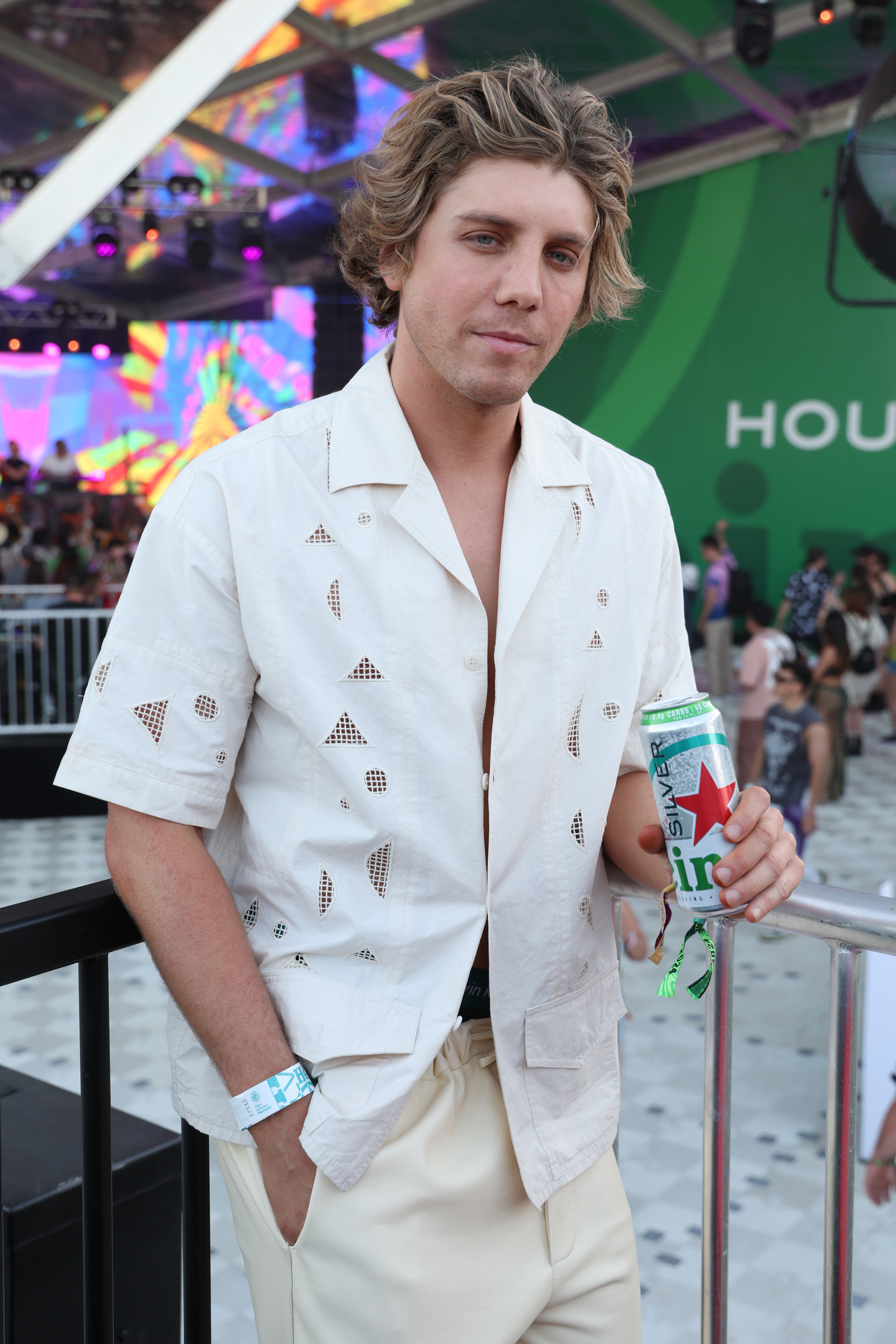 Man in a casual open shirt with pattern detail and pants holds a drink at an event