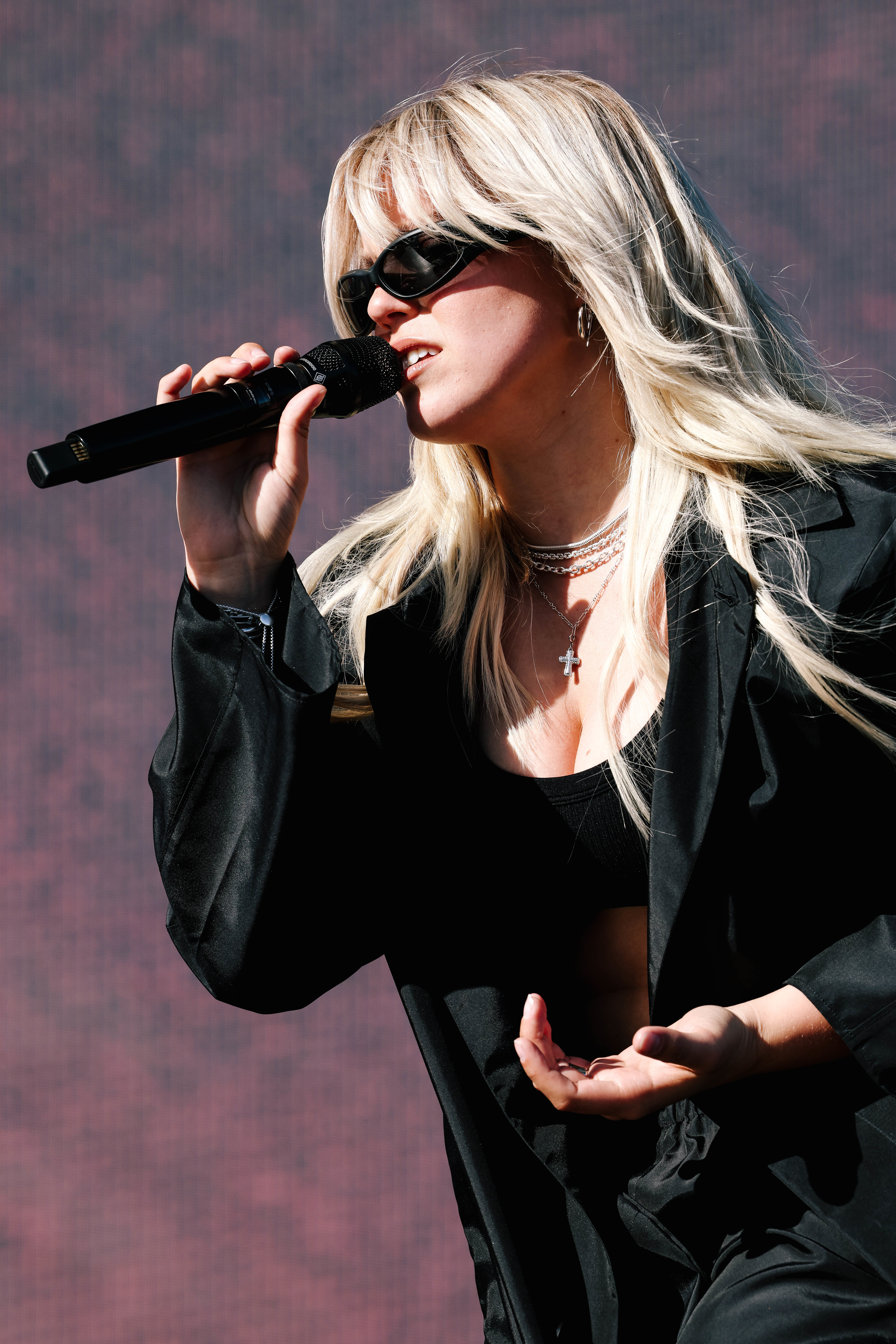 Singer with microphone performing on stage, wearing a black outfit with layered necklaces