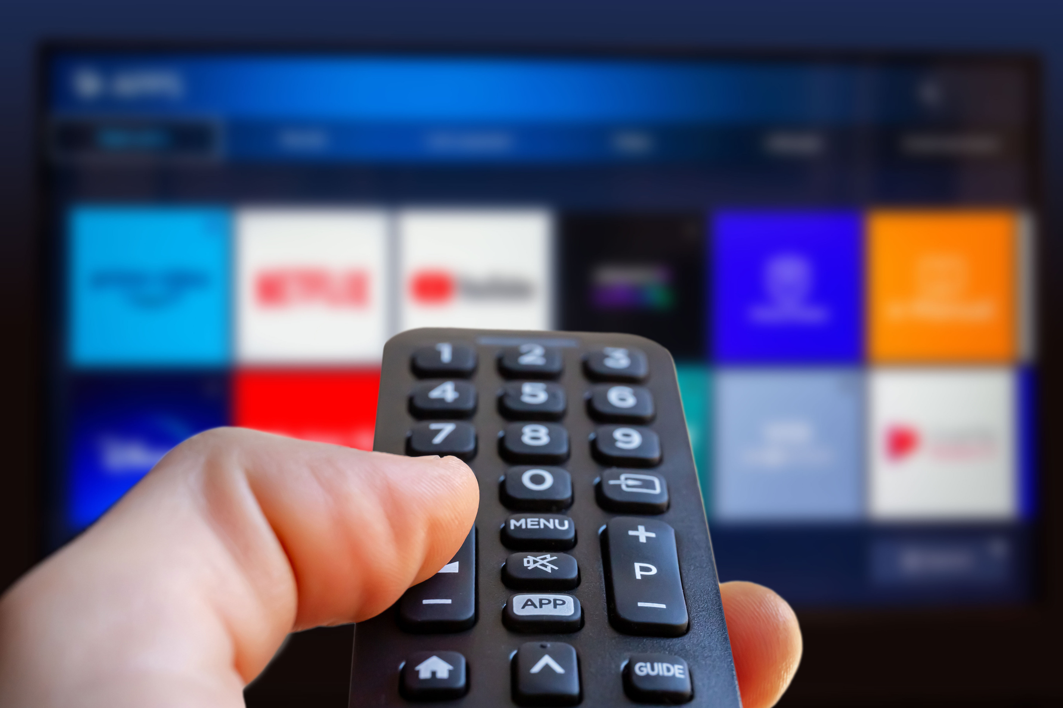 Hand holding a remote control, pointing at a blurred smart TV screen with app icons