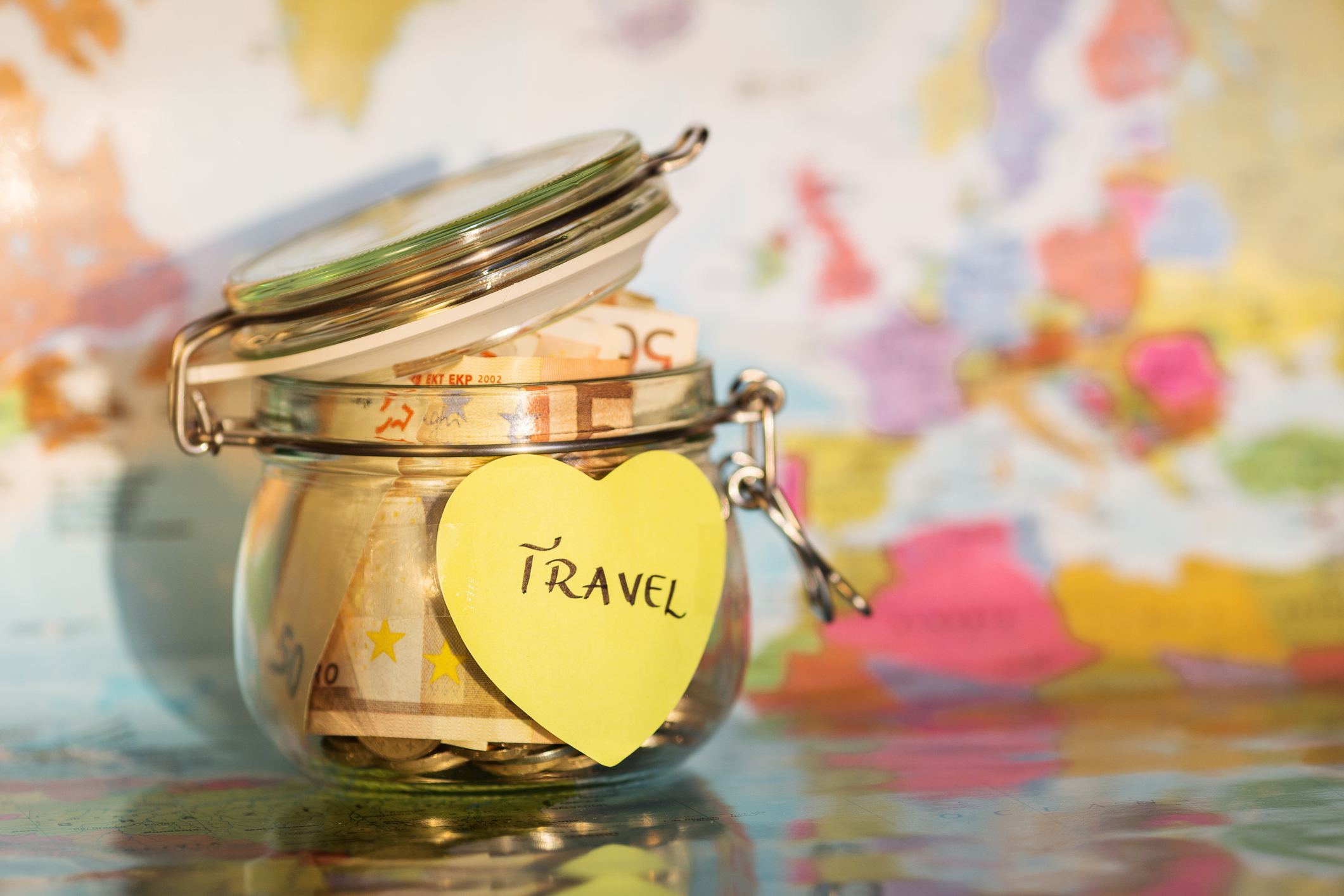 Glass jar with money on a map marked with a &#x27;Travel&#x27; sticker, symbolizing saving for trips