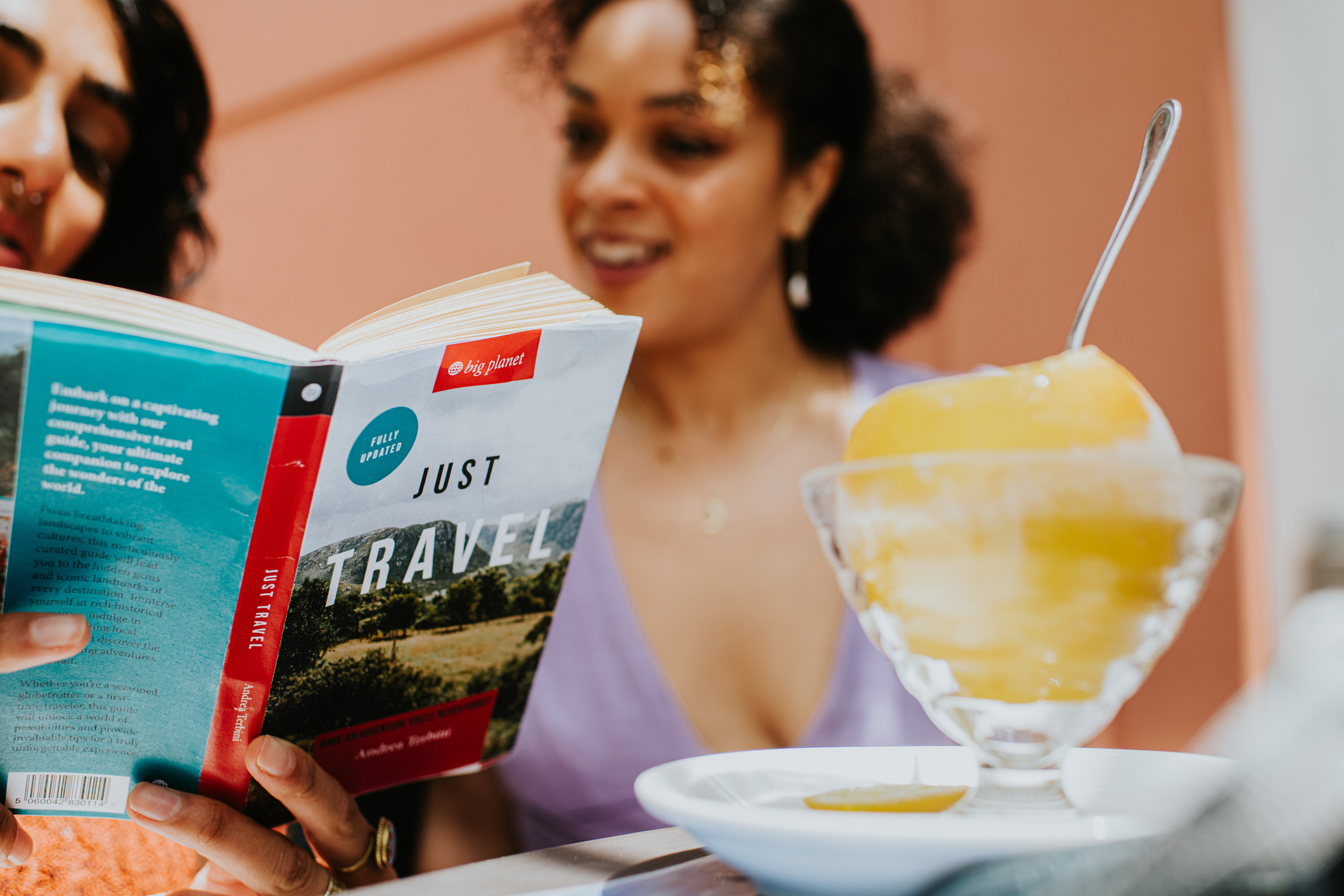 Woman reading a book titled &quot;Just Travel&quot; next to a bowl of dessert on a table