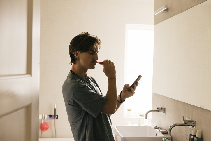 Man brushing his teeth while looking at his smartphone in a bathroom