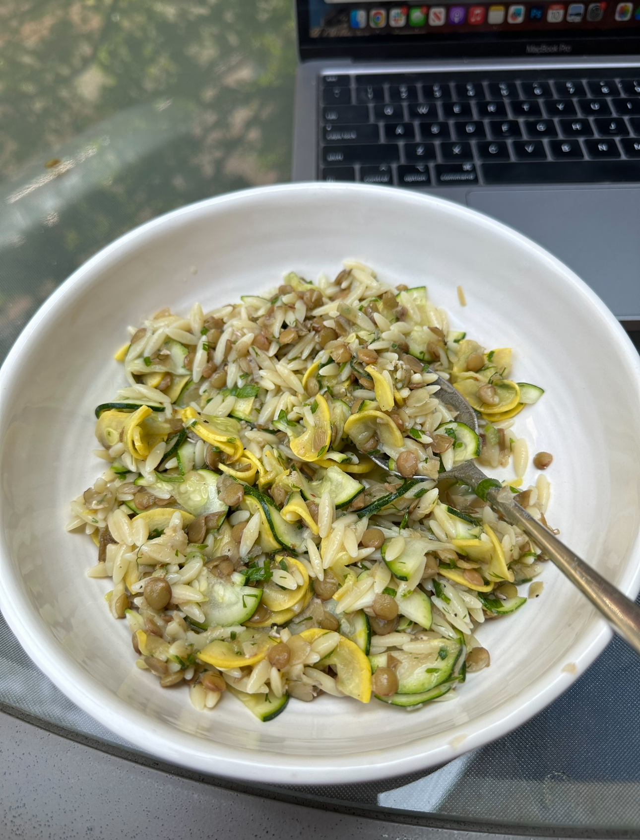 A bowl of zucchini and grain salad with a fork, placed on a table with a laptop in the background