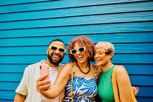 Three friends taking a selfie, smiling, with one holding a smartphone. They're wearing sunglasses and summer attire