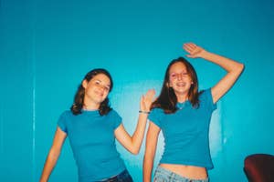Two individuals in matching blue tops, one waving, both smiling, in a room with a blue wall