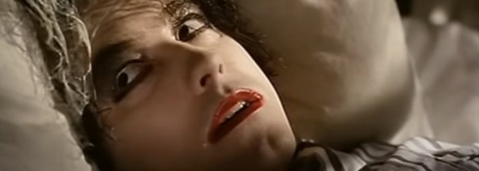 Robert Smith of The Cure in dramatic makeup lying down, appearing alarmed or in shock, in the Lullaby music video